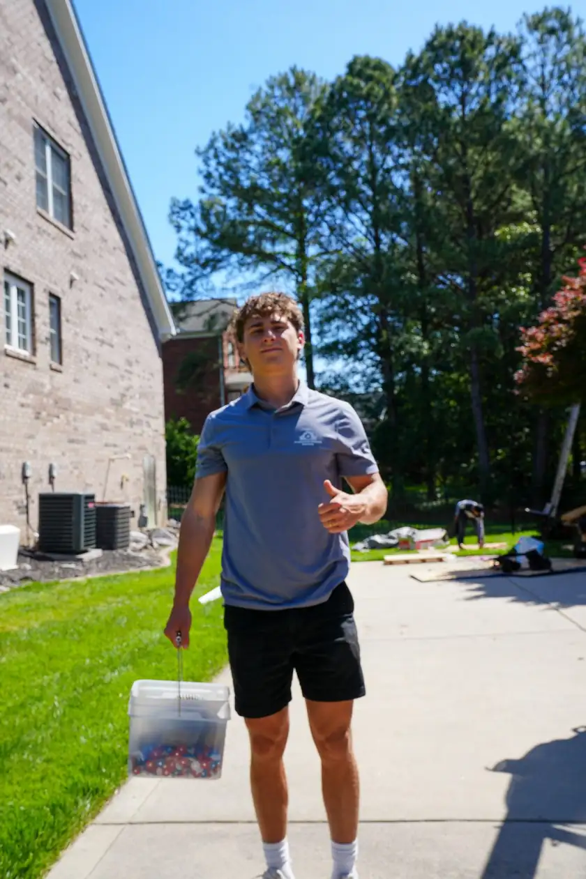 Charlotte Premier Roofing crew member carrying a tote of roofing fasteners up a driveway beside a brick home