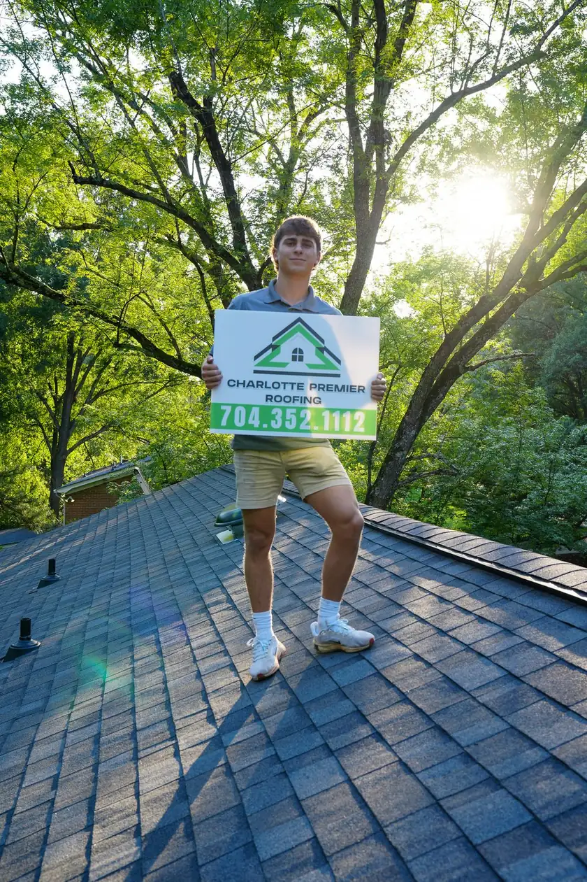 Charlotte Premier Roofing team member standing on a shingle roof holding the company yard sign beside vent pipes
