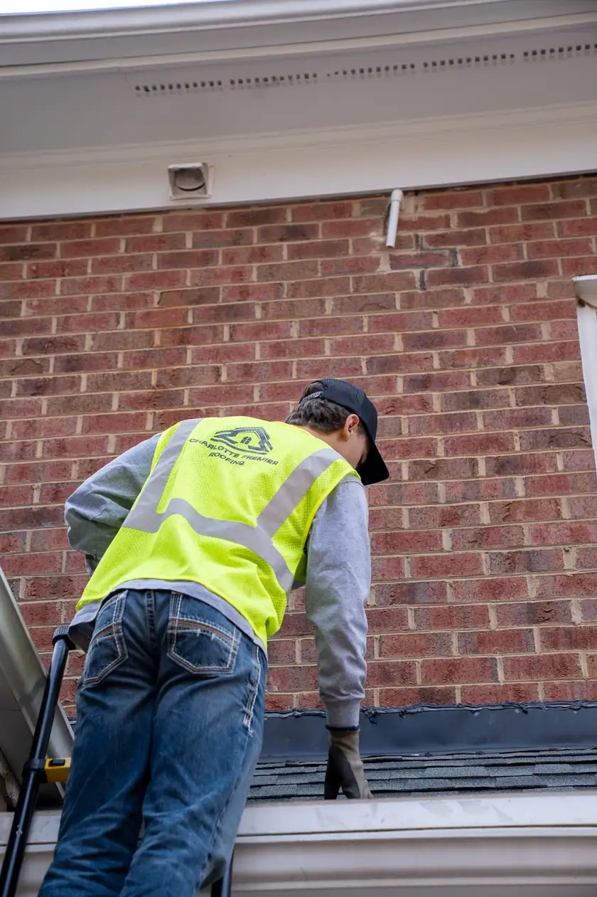 Charlotte Premier Roofing crew member on a ladder checking new flashing where a low roof meets a brick wall