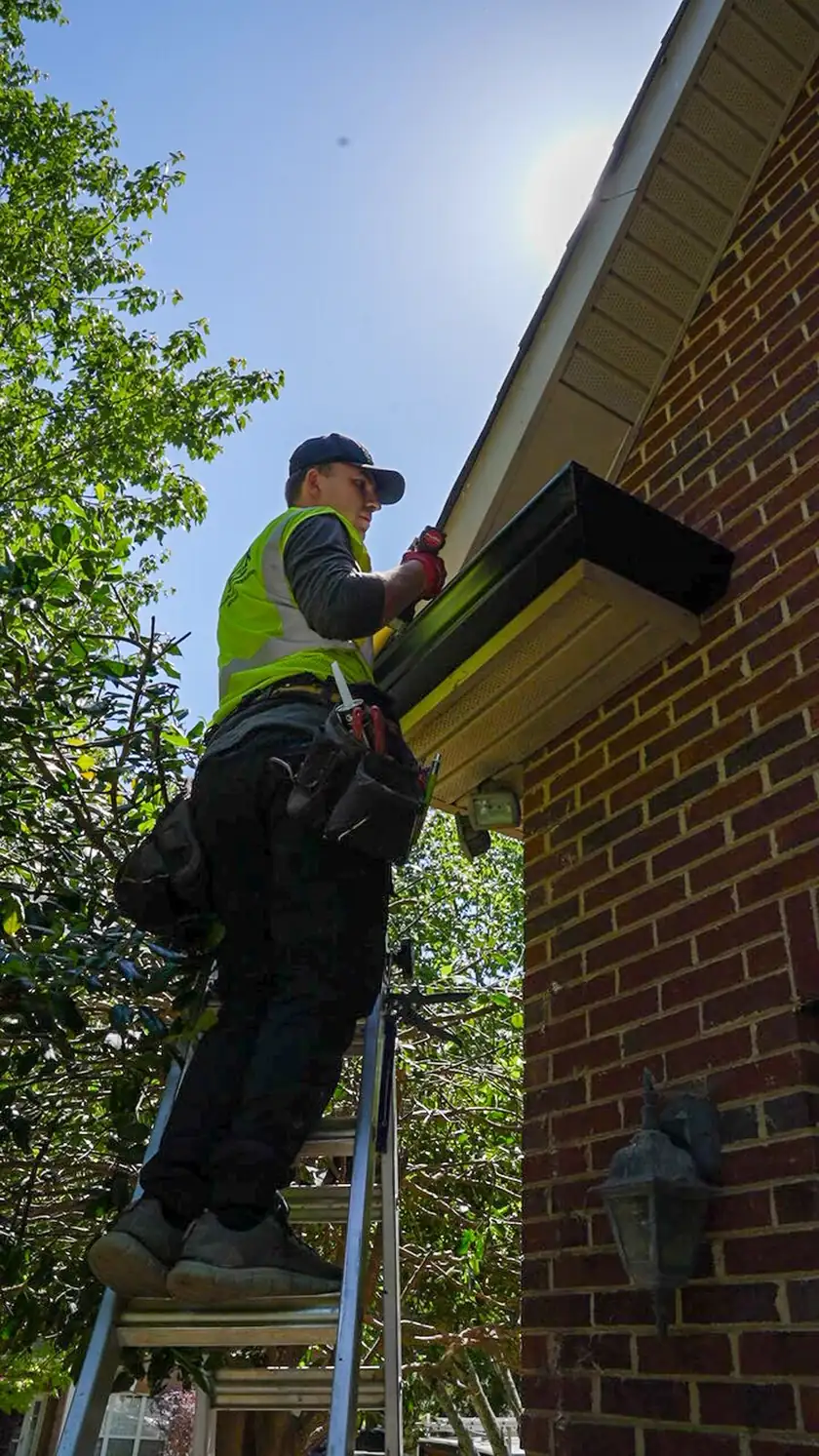 Charlotte Premier Roofing crew member on a step ladder fastening a new black gutter under the eave of a brick home