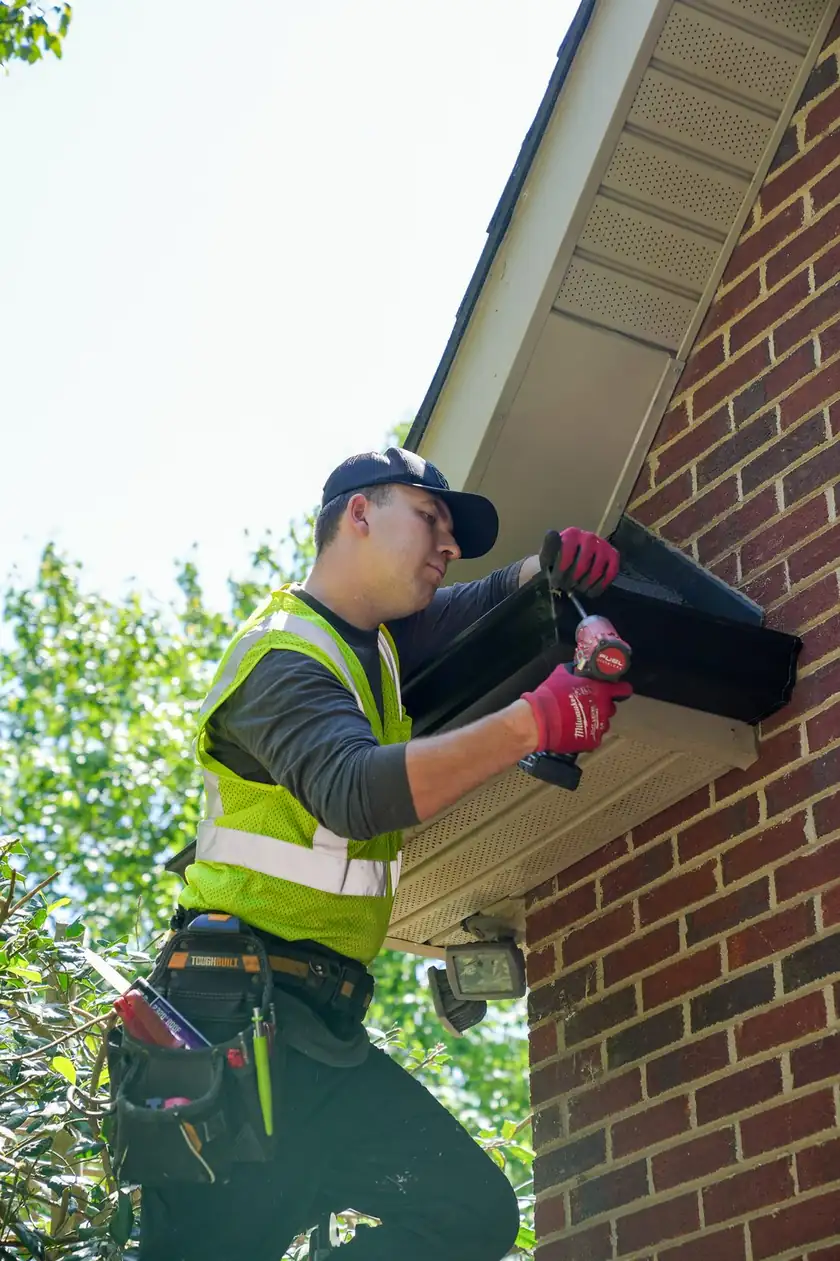 Charlotte Premier Roofing crew member fastening a new black gutter under the eave of a brick home in Weddington, NC