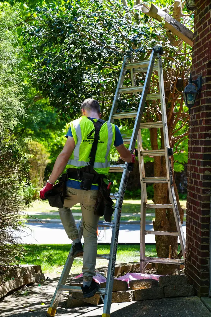 Charlotte Premier Roofing crew member stepping onto a ladder beside a brick wall, a second ladder set up behind him