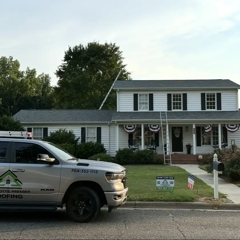 Charlotte Premier Roofing truck and yard sign outside a white two-story home with a ladder to the roof