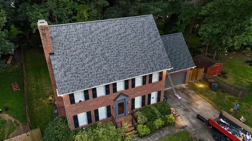 Drone view of a brick colonial with a new gray shingle roof, a brick chimney and trees behind