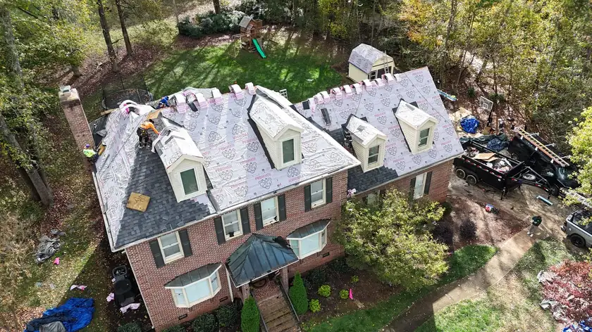 Drone view of a large brick home with dormers mid re-roof, shingle bundles on the ridges and trees all around